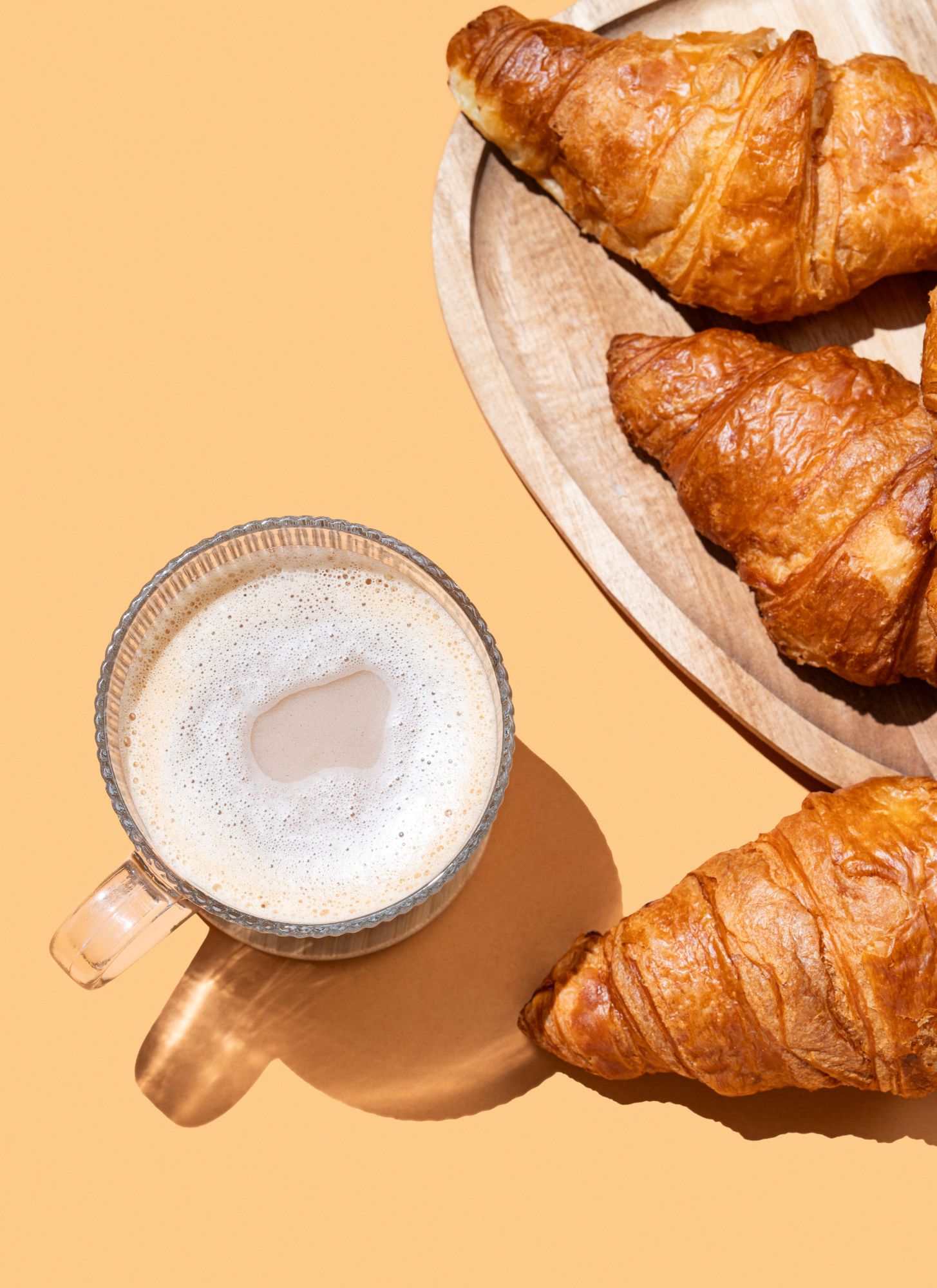 Ein Milchkaffee im Glas und Croissants auf einem Holzbrett, von oben fotografiert.